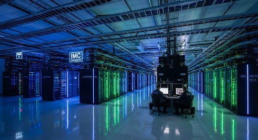 A high-tech data center with glowing blue and green server racks. The image is captured with a wide-angle lens, showing the vast scale of the facility. The floor is reflective, and the ceiling has clean cable management. The mood is secure, advanced, and highly technical. Corporate technology aesthetic.